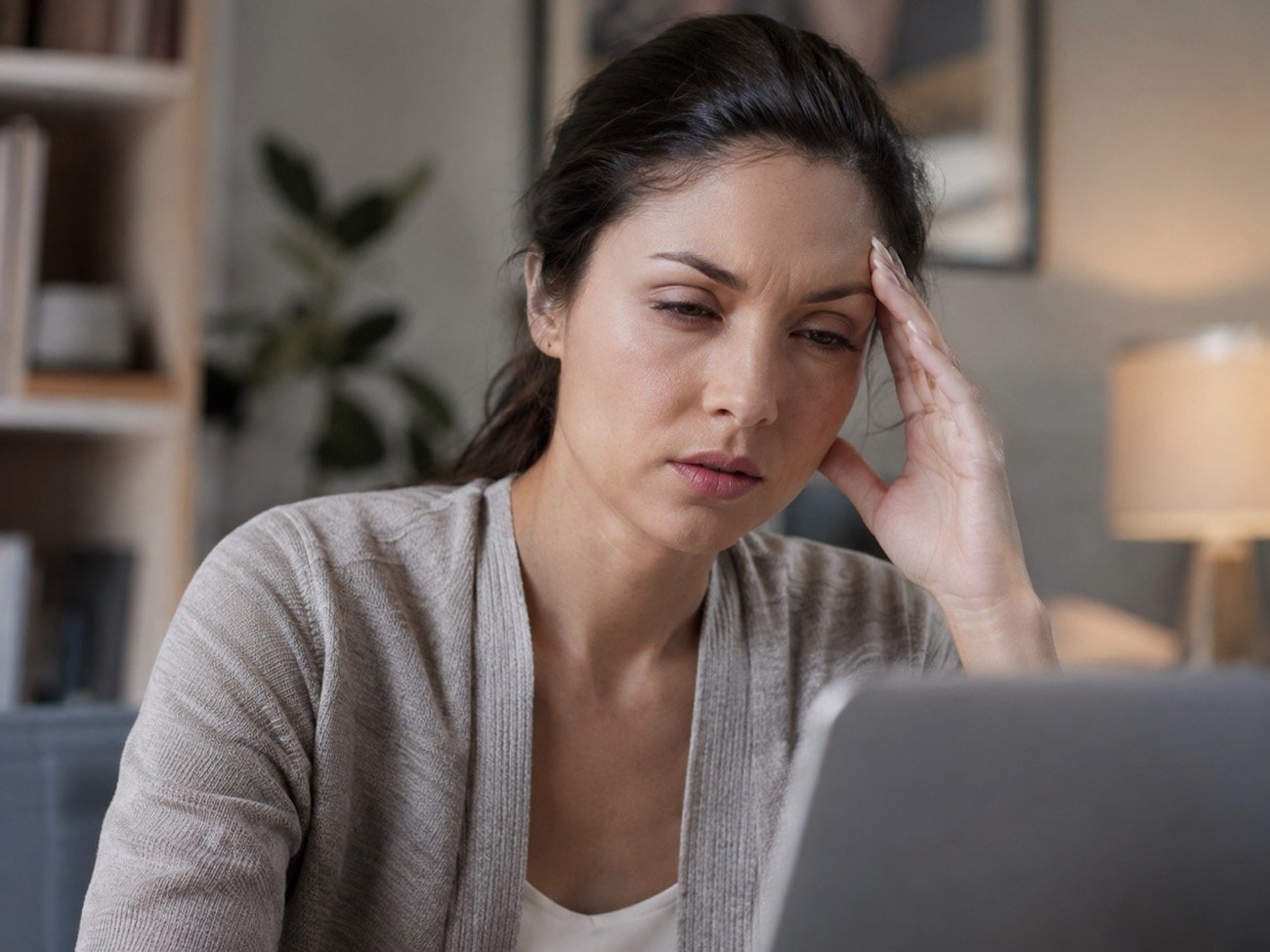 A woman at a laptop pressing her fingers to her temple, eyes tired and brow furrowed
